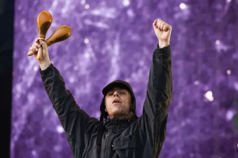 Getty Images Liam Gallagher raises his hands into the air during a concert in 2024. In his right hand, he is holding a pair of maracas.