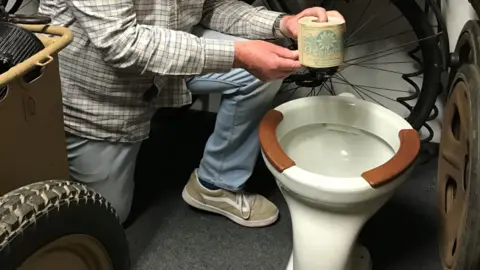 Graeme Delanoe kneels holding a roll of occupation-era toilet roll above a toilet bowl with is white porcelain with two brown wood panels around the top of the bowl. Behind are various WW2 items stacked on shelves.