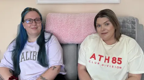 Kate Bradbrook/BBC A woman with long purple and brown hair and mauve T-shirt sits on a sofa next to another woman with shoulder-length light brown hair and a cream T-shirt.