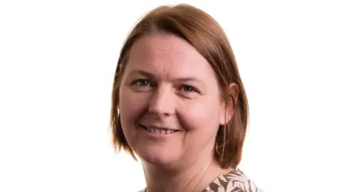 Claire Shrewsbury/WRAP A woman with light brown hair smiles at the camera. She wears a brown top with white leaves on. Head and shoulders on a white background.