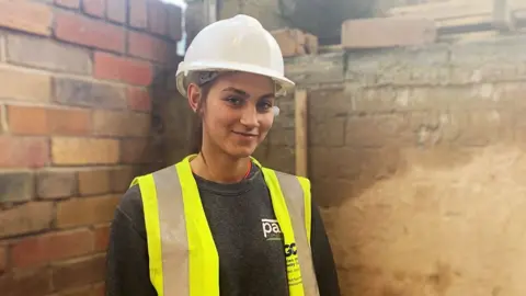 A young woman with dark hair in a white hard hat and yellow high visibility bib over a black jumper stands in front of a brick wall