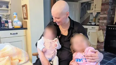 A mother, who has lost her hair as a result of chemotherapy, cradles an infant girl. She is sitting in an armchair a kitchen. Next to them is a young girl. She is wearing a pink long-sleeved t-shirt. The faces of the children have been blurred.