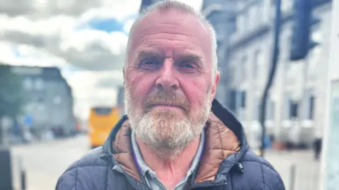 Man - Ron Constable - with grey beard, looking at camera, with busy street in background.