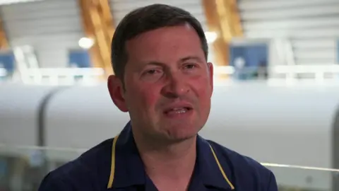 A man sits speaking wearing a navy nurse's uniform in a hospital building. 