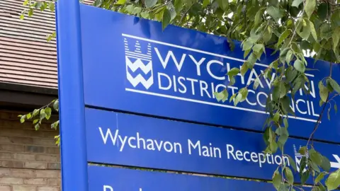 A blue sign outside a building, partly covered by leaves. The sign reads Wychavon District Council. Another sign with the words Wychavon Main Reception is directly underneath.