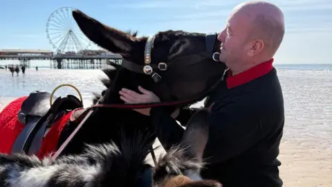 Mark, wearing a black jumper and red polo shirt underneath, turns towards a brown donkey that is resting its head on his shoulder. They are on Blackpool beach with the pier and Big Wheel visible in the background beyond the sand and sea.