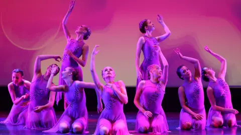 A group of dancers, in a dance pose, all in lilac colours. Two are standing up and the rest are kneeling down on the floor, mostly with one arm raised above their heads.