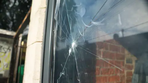 Close up photograph of a smashed window. There is clear film over the top of the crack. Red brick wall can be seen through the window to the inside of the coffee shop. 
