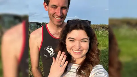 Chloe Morley Tom and Chloe standing next to each other smiling in a field. She's holding up her hand to show off her engagement ring