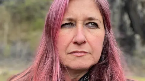 Helen looks into the distance, away from the camera, with a serious expression. She has long dyed pink hair, and is standing outside with blurred greenery behind her. 