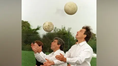 PA Media Former prime minister Tony Blair takes part in a ball header competition with former Newcastle United boss Kevin Keegan. Both men have a white football above their heads that they are looking at. They have their arms stretched out in front of them. They are both wearing white shirts. 