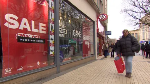 BBC A shopper passes a shop window with red sale, 70% off signs on a high street. There are other people walking in the background, some with shopping bags and a Halifax sign. There are tree branches on the right of the picture. 