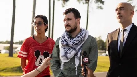 Mahmoud Khalil listens to a reporter question  after he is released from immigration custody in Jena, Louisiana. He is flanked by two people: a man in a black suit and woman wearing a red Mickey Mouse shirt.