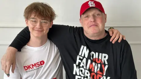 Karol Sadlisz and his dad Gregg Sadlisz , who have their arms around each other. Karol has mousy coloured hair, and is wearing glasses and jeans. Gregg is wearing a cap and black jeans. Both are wearing t-shirts relating to blood cancer; Karol's is white and Gregg's is black.