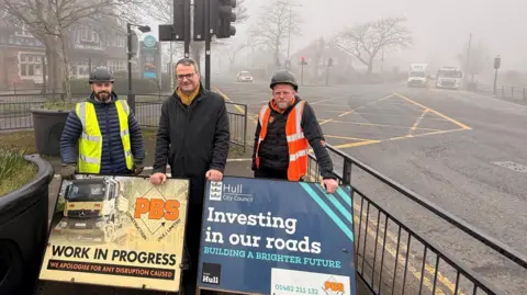 Three men standing behind signs on the footpath beside a large road junction on a foggy day. The man in the centre has short hair and wears a smart coat, glasses and a mustard-coloured scarf. He is flanked by two men wearing black hard hats, black coats and hi-vis jackets. The sign on the left includes a picture of a truck and words reading: "PBS: Work in progress, we apologise for any disruption caused". The sign on the right has a Hull City Council logo and words reading: "Investing in our roads, building a brighter future". Cars and trucks are waiting at the junction, which includes a yellow box pattern. A large pub called The National is on the left.