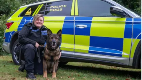 Northamptonshire Police PC Samantha Clark, in her police uniform, smiles as she kneels next to Olly, a German Shepherd, alongside a police vehicle. 