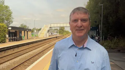 Richard Stow has short grey hair and is wearing a light blue, striped shirt. He is stood on a railway station platform. There is a metal footbridge and a brick station office behind him.
