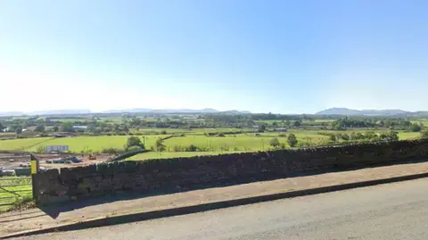 A Google Street View screenshot of the land where the housing development is proposed, seen from the A6. Green fields stretch out behind a stone wall with traditional farm gate. There are pockets of trees and, in the far distance, the tops of mountains rise.
