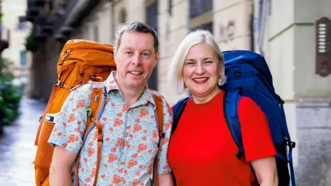 BBC/Studio Lambert In-laws Mark and Margo with their backpacks on, smiling at the camera