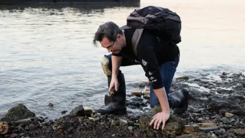 Mudlark Jason Sandy wearing a backpack is bent over and holding a trowel as he searches the shoreline of the River Thames for historical items