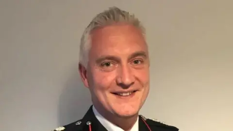 West Midlands Fire Service Ben Brook has short grey hair and he is smiling at the camera. He is wearing full dress Fire Service uniform.