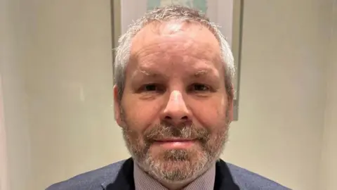 Sean McNamee. He has short silver hair and beard, wearing a dark blue blazer and check shirt. Behind him is a wall with a photo frame on it.