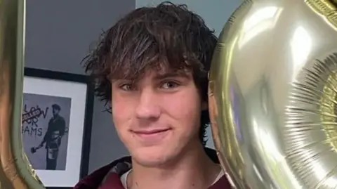 George is holding two gold balloons close to his head and smiling. He is inside a house and a picture can be seen in the wall behind him. He has short, brown hair with a fringe.