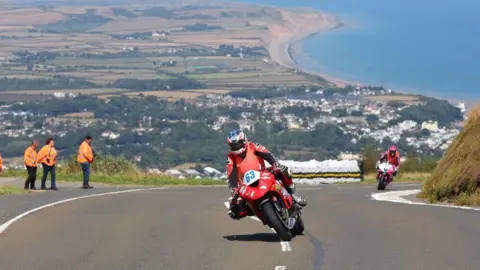 Dave Kneen Photography Two bikes on climb up the Mountain Road, one is red and is followed by another that is pink and there are marshals in orange jackets on the left. There is a picturesque scene behind them of green fields and the blue sea of the sweep of Ramsey Bay. 