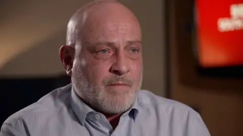 John Pickering wears a blue button shirt while sat in a dark room during an interview being filmed for television. He is bald with a white beard and is looking to the right out of frame. The background is out of focus. "BBC South Today" branding can vaguely be made out on a tv screen on the wall. 