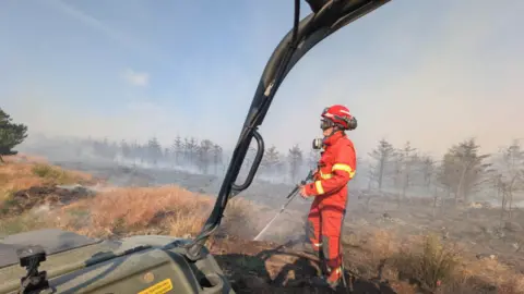 A firefighter in bright red protective clothing stands in a smoke-filled heathland, spraying water onto scorched ground after a fire. The scene is viewed from inside an off-road vehicle, with part of the vehicle frame visible in the foreground. Burnt vegetation and a line of smoke-shrouded trees stretch across the background under a pale blue sky.