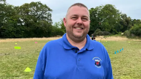 Nathaen, who has short brown hair and stubble and wearing a blue polo shirt, is smiling at the camera. He is stood on a grass football pitch with yellow and blue cones and small goals behind him.