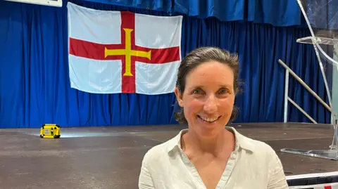 Deputy Lindsay de Sausmarez stands in front of a stage which has a Guernsey flag in the background. She is smiling.