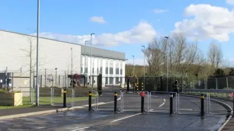 A grey industrial building behind a number of large bollards and fencing. 