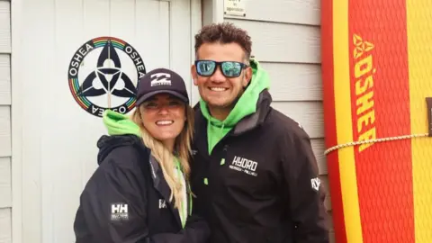 Andy Johnson Photograph of Andy Johnson (right) and Danni (left). They both wear black rain coats and green neon hooded jackets underneath. Andy wears sunglasses and has brown hair. Danni wears a black cap and has long blonde hair and smiles at the camera. 