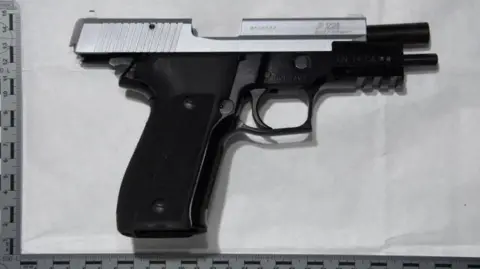 Avon and Somerset Police A 9mm pistol laying on a sheet of paper on a table, taken from above. The barrel has been cocked open and is empty. On either side of the gun is a measuring ruler.