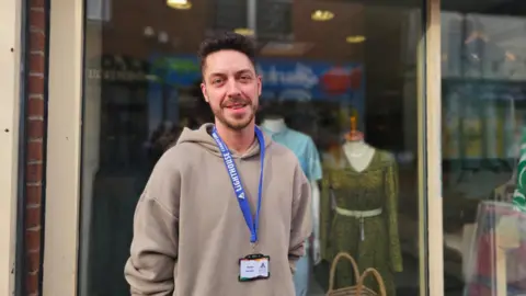 Steven Caulton has dark brown short hair and is wearing a beige hoodie. He is in front of his shop front and has a blue lanyard around his neck.