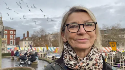 Robby West/BBC Sandi Lowe is on the right of the frame. She has shoulder length blonde hair and is wearing black glasses and a leopard print scarf. Behind her is Norwich Market and pigeons flying.