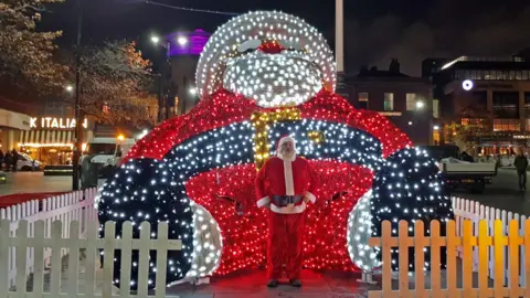 Derby City Council The UK's largest light-up Santa installation behind a man who is dressed up as Santa Claus himself
