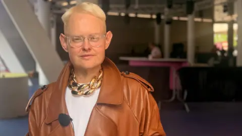 Michael Hopkinson, who is wearing a brown leather jacket, gold chain and white T-shirt, is staring directly at the camera. 