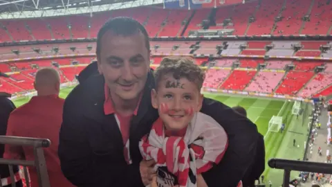 Brian Soulsby and his son Ben are standing in the South Stand at Wembley Stadium ahead of Sunderland's match with Sheffield United. The duo are both dressed in red and white Sunderland shirts. 