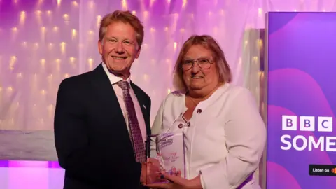 Simon Parkin wearing a suit and tie, shaking hands with Cath Morrison, who is wearing a white blouse and large glasses. They are both looking at the camera and smiling, while holding her glass award. 