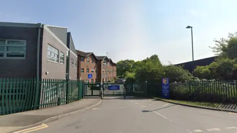 Google A Google Street View screenshot of the outside of a school. It has dark green railings and double gates closed over the road. A blue sign next to the road says the name of the school and there are several brick buildings behind the railings.