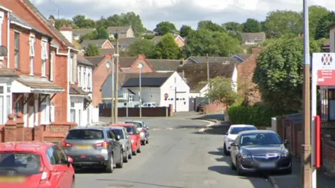 A Google streetview image of a residential street. Cars are parked on both sides of the road and there is a bus stop.