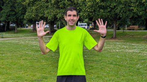 Picture shows Kieron standing in a park holding up ten fingers, wearing running clothes. 
