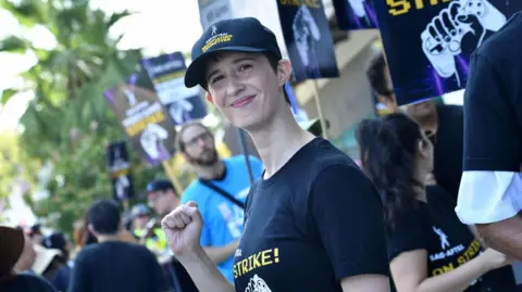 A woman with short black hair wears a black baseball cap and t-shirt with matching Video Game Actors' strike logos, with text and a picture of a hand holding a control pad.