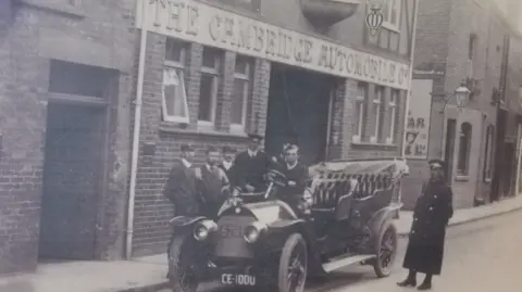 Museum of Cambridge 7 men standing by a 1900s car outside a large building with The Cambridge Automobile Company written on the sign. Two of the men wearing police uniforms
