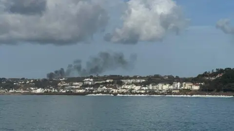 A view from afar of houses and roads in the distance, with a plume of black smoke coming from the left area. The sea is in the forefront.