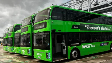 Five green double decker buses with "west bus" in white text are parked in a depot. 