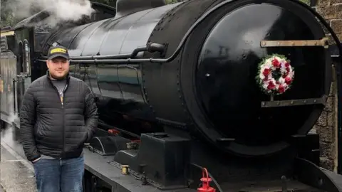 A man in a black zip-up puffer jacket stood next to a black steam train with a flower wreath attached to the front.