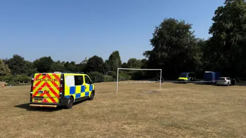 A field with a football goal in the middle. There is police tape across the field and a police van to the left. To the right is three other vehicles.